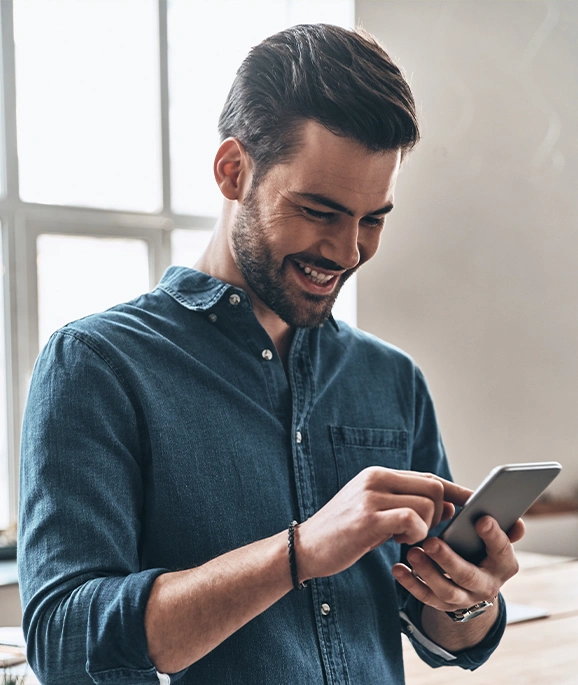 Um homem com uma camisa jeans sorri enquanto usa um smartphone em um ambiente interno, perto de uma janela iluminada pelo sol.