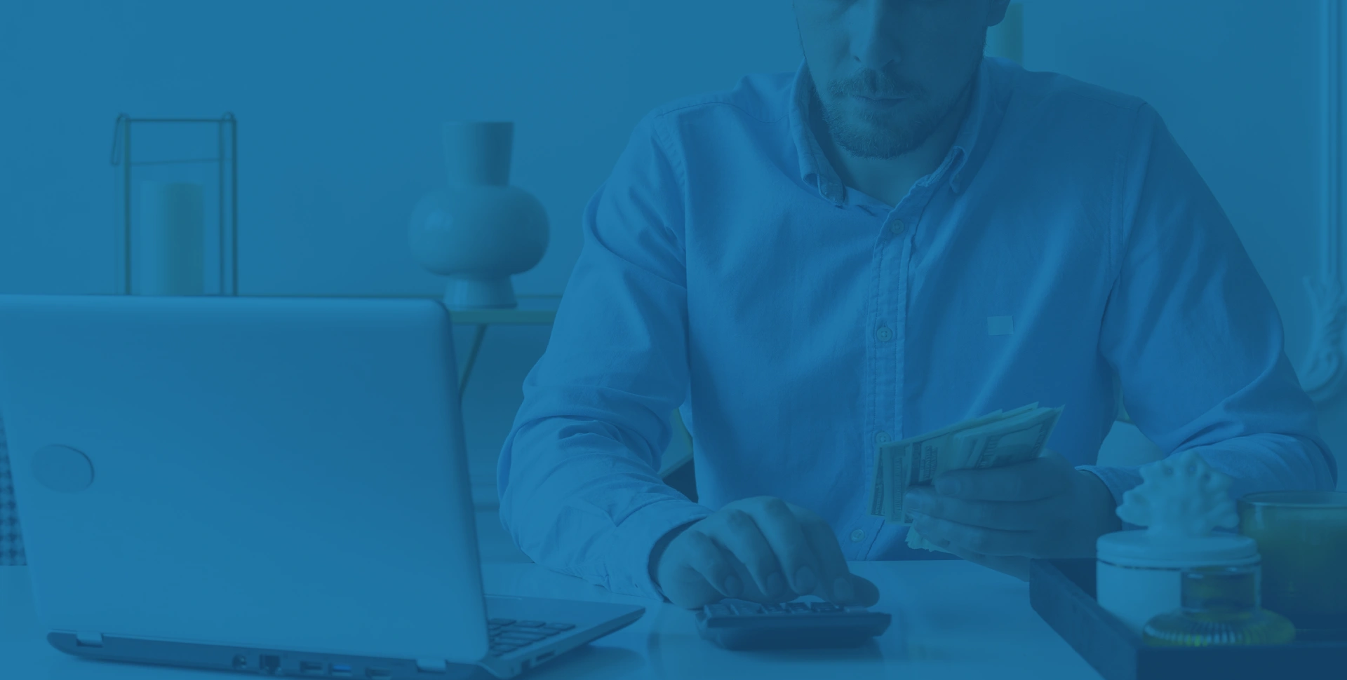 Um homem está sentado em uma mesa usando uma calculadora, segurando dinheiro em uma das mãos, com um laptop aberto e uma caneca por perto.